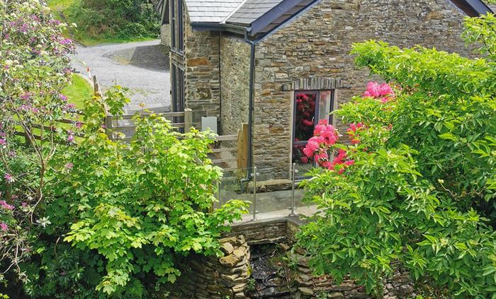 A charming stone cottage nestled among flowering shrubs and trees, with a babbling stream flowing below the patio.